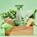 Sustainable home cleaning products featuring reusable spray bottles and natural brushes arranged in wooden crate with green ferns